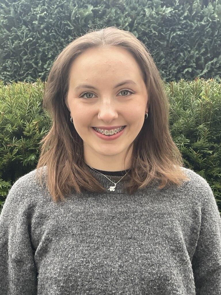 Junge Frau mit Brille und lockigem, braunem Haar in grauem Pullover vor grünem Hintergrund.