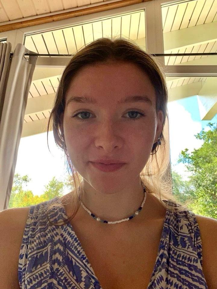 Porträt einer Person mit blauer Bekleidung und Halskette, sitzend vor einem Fenster mit Tageslicht und grünen Pflanzen im Hintergrund.