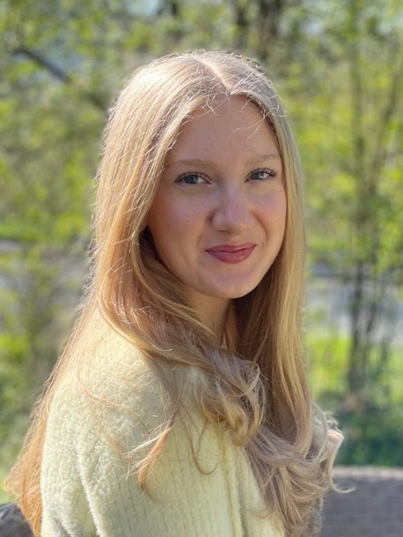 L&auml;chelnde junge Frau mit langen, blonden Haaren in einem hellen Pullover, vor einer gr&uuml;nen Naturkulisse.