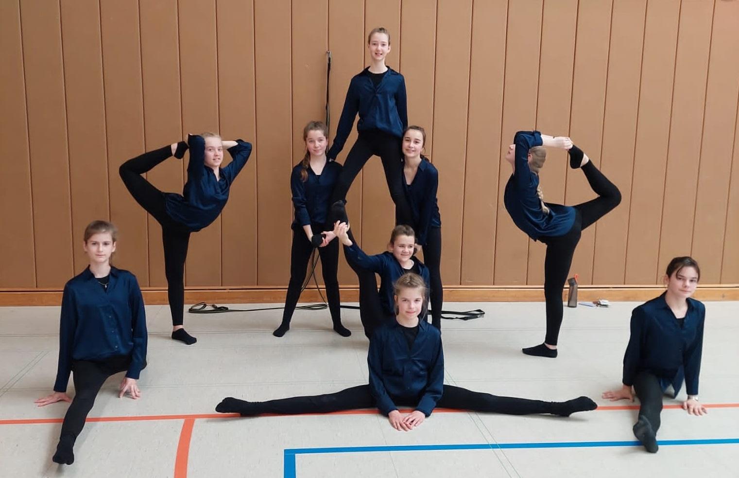 Gruppe von acht T&auml;nzern in blauen Oberteilen, in verschiedenen Posen auf einer Sporthalle.