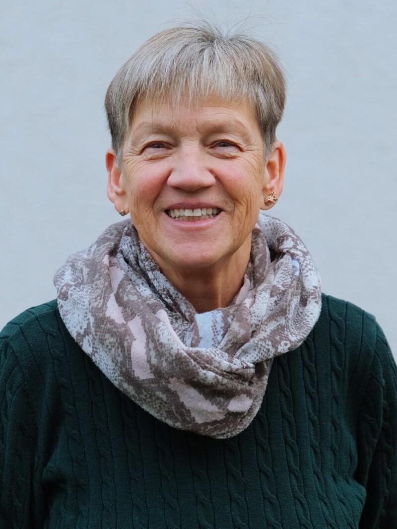 Lächelnde Frau mit kurzem, grauem Haar, grünem Pullover und gemustertem Schal vor heller Wand.