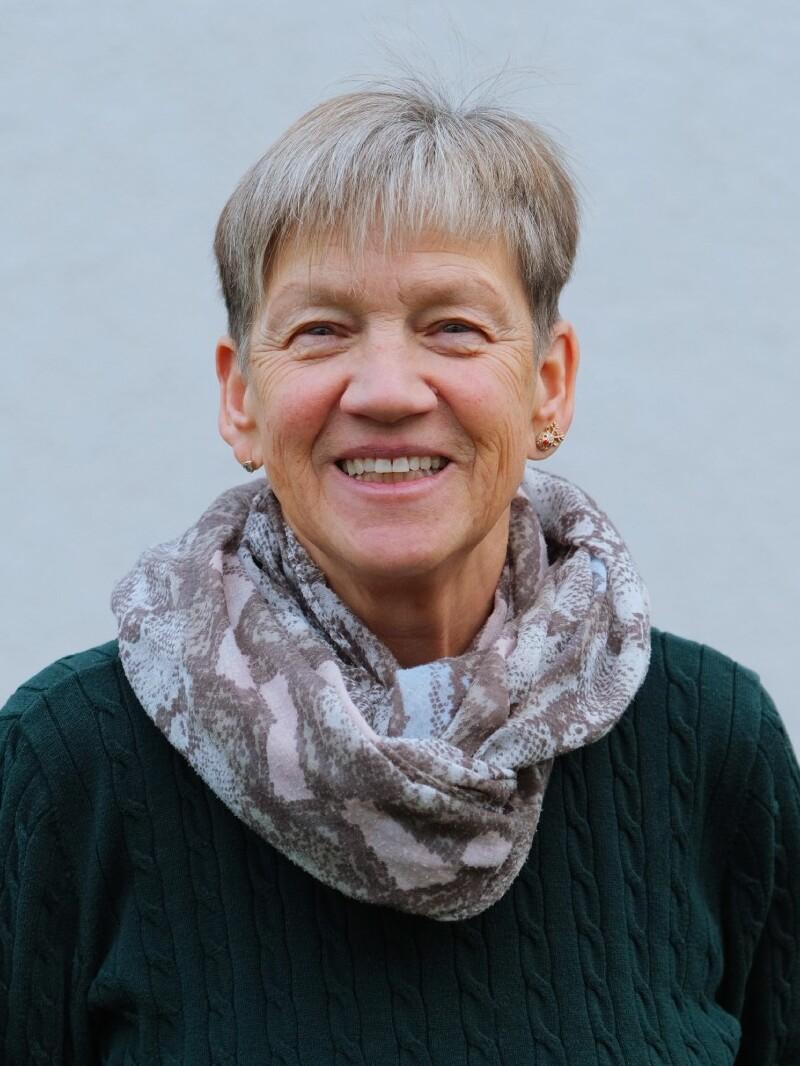 Lächelnde Frau mit kurzem, grauem Haar, grünem Pullover und gemustertem Schal vor heller Wand.