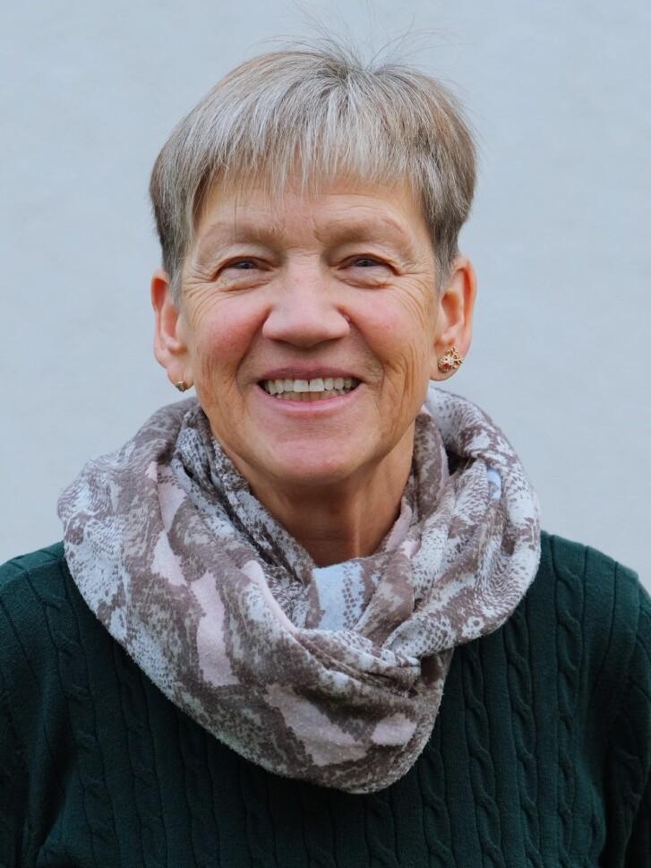 Lächelnde Frau mit kurzem, grauem Haar, grünem Pullover und gemustertem Schal vor heller Wand.