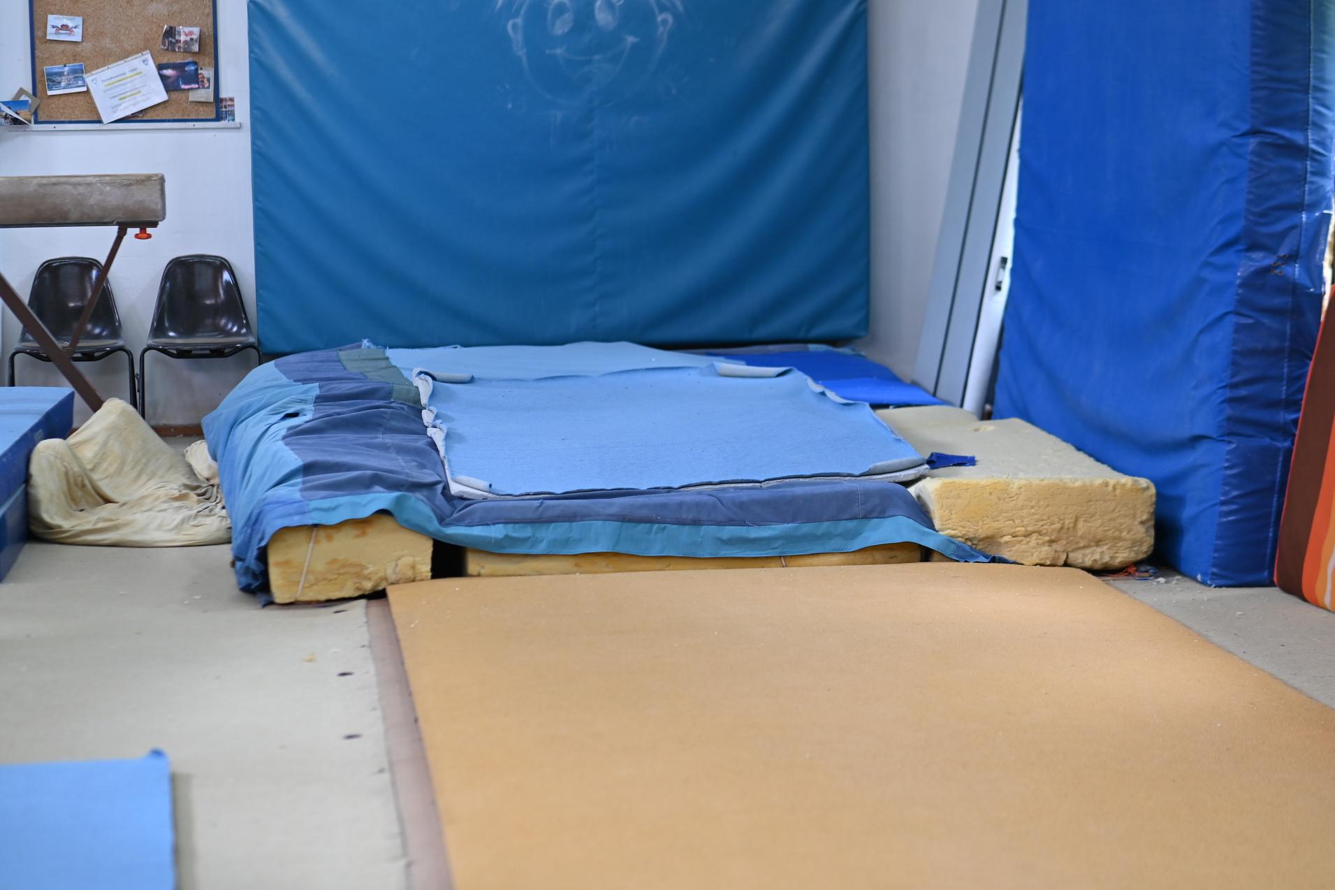 Kleine Turnarena mit gestapelten Matten, blauer Polsterung und St&uuml;hlen im Hintergrund.