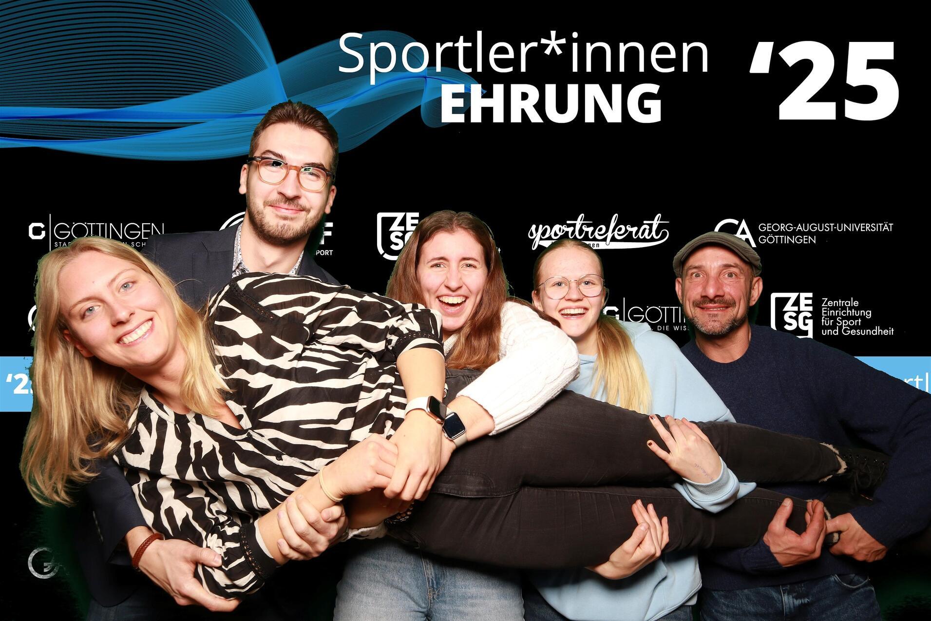 Gruppe von vier Personen, die eine Frau hoch halten, vor einem Hintergrund mit der Aufschrift Sportehrungen '25