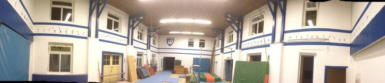 Panoramablick auf einen Trainingsraum mit blauen W&auml;nden, Matten und Holzrahmen in einem Gymnastikbereich.