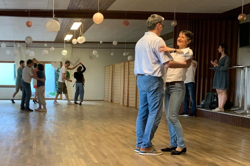 Tanzpaar im Vordergrund umarmt sich auf einer Holzfl&auml;che, w&auml;hrend im Hintergrund weitere Paare tanzen.