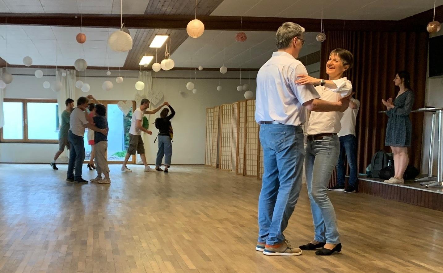 Tanzpaar im Vordergrund umarmt sich auf einer Holzfläche, während im Hintergrund weitere Paare tanzen.