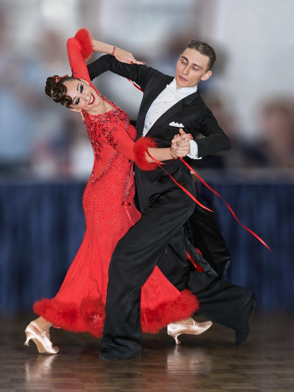 Tanzpaar in eleganter Tanzhaltung, Frau in rotem Kleid mit R&uuml;schen, Mann in schwarzem Anzug, vor verschwommenem Hintergrund.