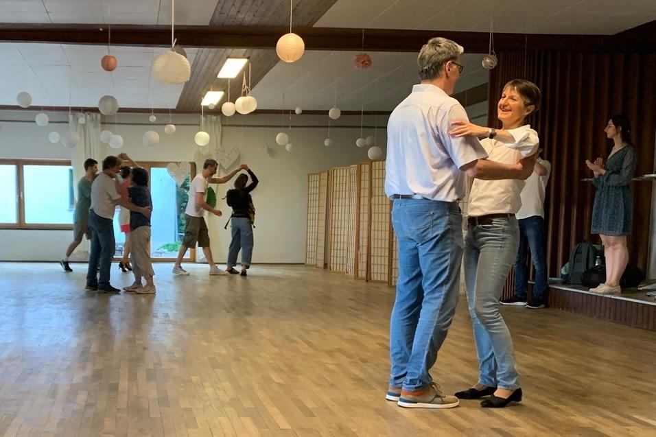 Tanzpaar im Vordergrund umarmt sich auf einer Holzfläche, während im Hintergrund weitere Paare tanzen.