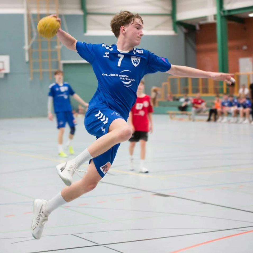 Aktiver Handballspieler in blauer Sportkleidung springt und wirft einen Ball in der Sporthalle während eines Spiels.