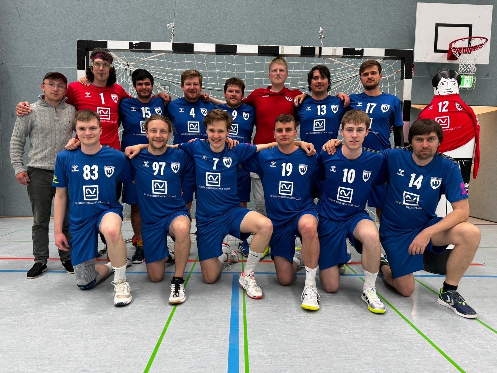 Gruppenfoto einer Handballmannschaft in blauen Trikots, posierend vor dem Tor in einer Sporthalle.