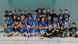 Gruppe von Kindern in blauen und schwarzen T-Shirts posiert l&auml;chelnd in einer Sporthalle vor einem Trainer.