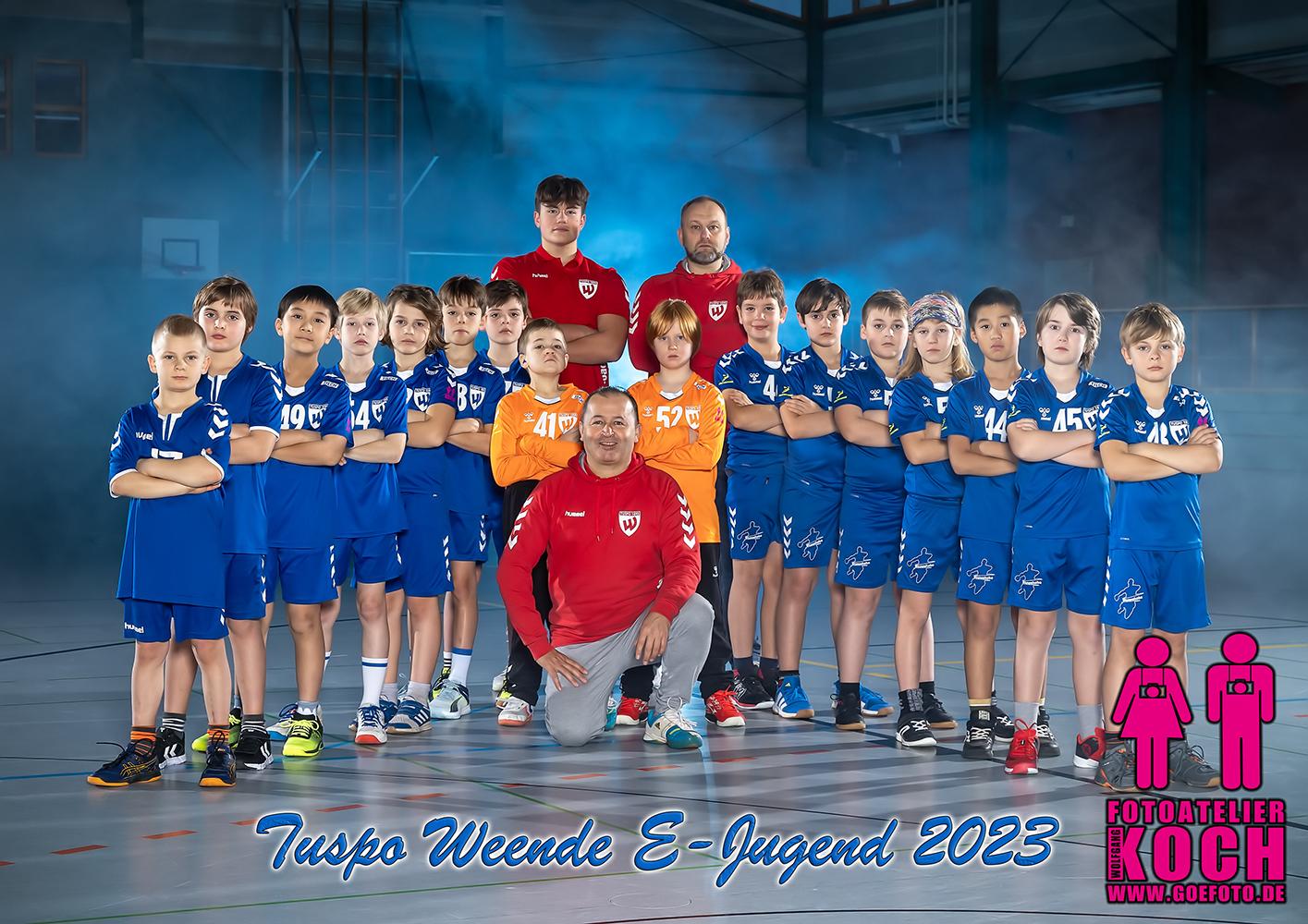 Gruppenfoto der E-Jugend von Tuspo Weende 2023 mit Spielern in blauen Trikots und Trainern in rotem Outfit in einer Halle.