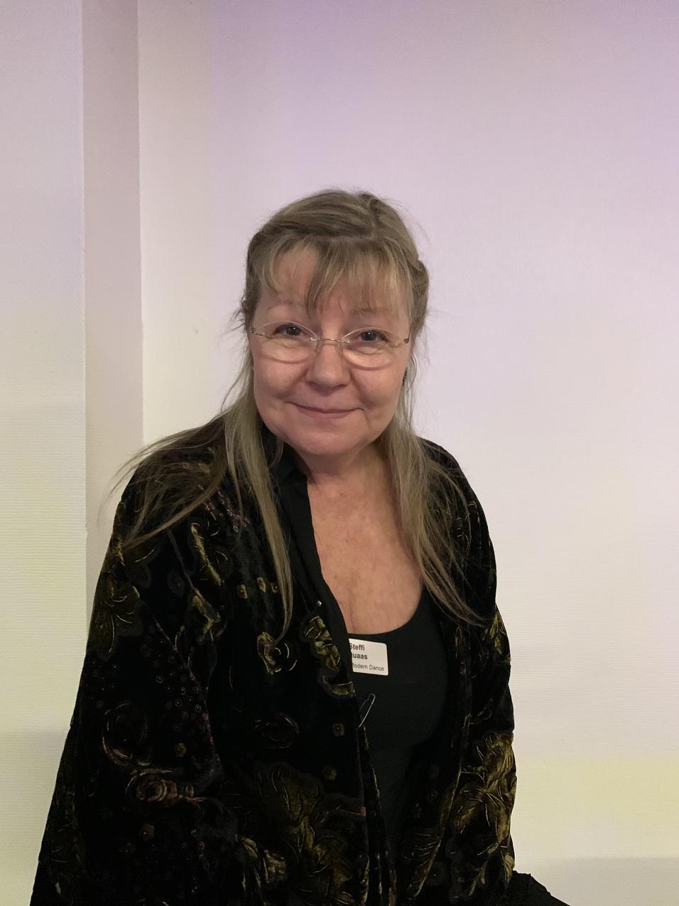 Porträt einer Frau mit langen Haaren, die eine dunkle Jacke trägt und lächelt, vor einem hellen Hintergrund.