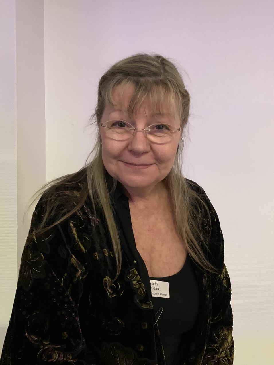 Porträt einer Frau mit langen Haaren, die eine dunkle Jacke trägt und lächelt, vor einem hellen Hintergrund.