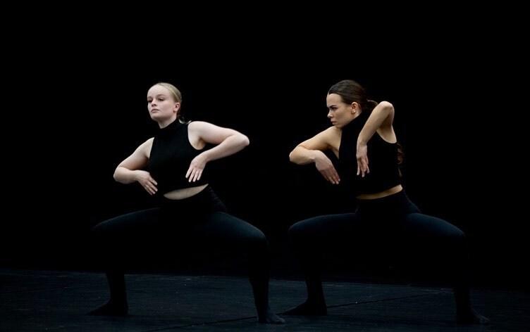 Zwei Tänzerinnen in Schwarz führen synchronisierte Bewegungen vor einem dunklen Hintergrund aus.