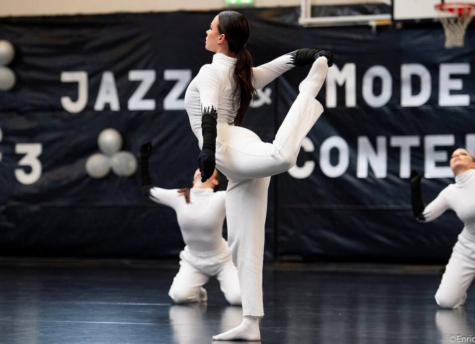 Tänzerin in weißem Outfit führt eine hohe Beinschwingung auf einer Bühne mit dunklem Hintergrund durch.
