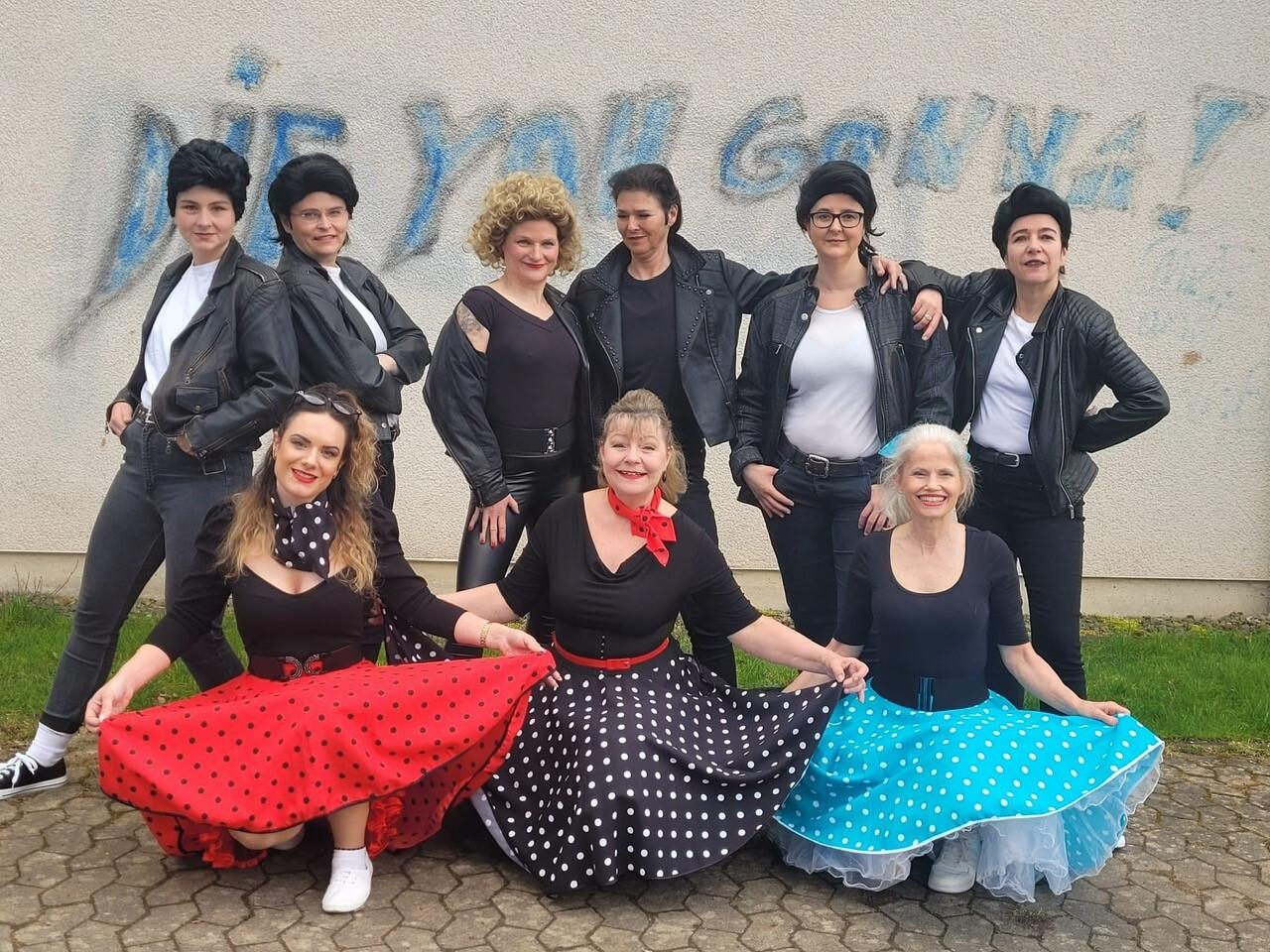 Gruppe von Frauen in Rock'n'Roll-Kleidung posiert vor einer Wand mit dem Graffiti &bdquo;DIE YOU GONNA!&ldquo; im Hintergrund.
