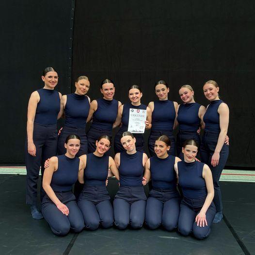 Gruppe von zw&ouml;lf T&auml;nzerinnen in blauen Kost&uuml;men, die stolz eine Urkunde in der Mitte halten.