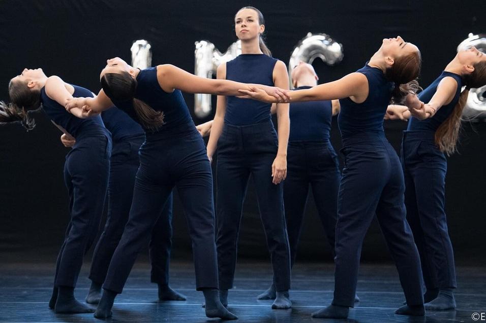 Gruppe von Tänzerinnen in blauen Outfits, die synchron eine dramatische Pose auf einer Bühne einnehmen.