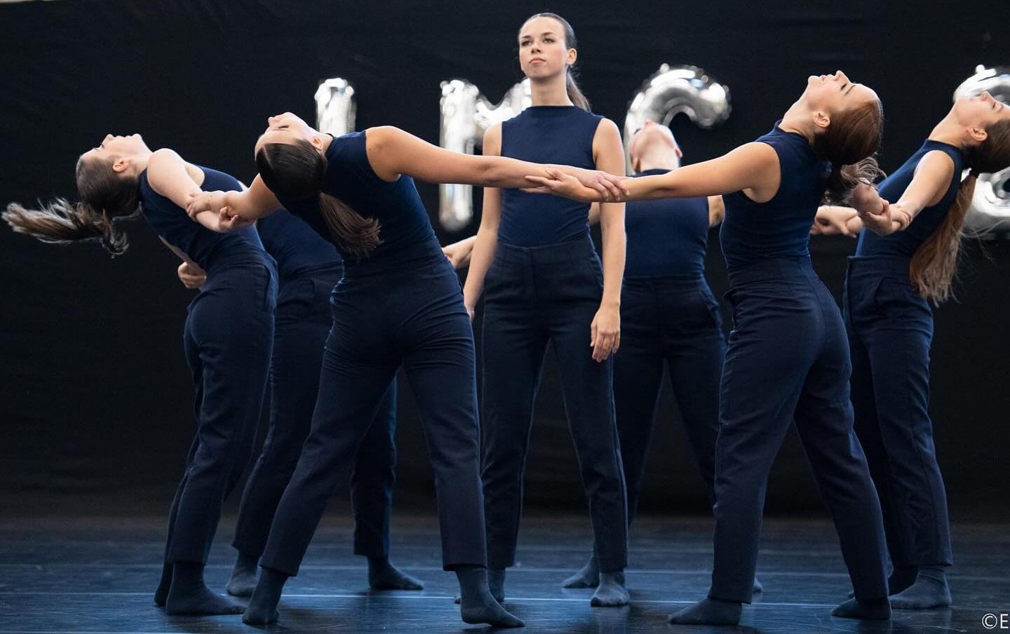 Gruppe von Tänzerinnen in blauen Outfits, die synchron eine dramatische Pose auf einer Bühne einnehmen.