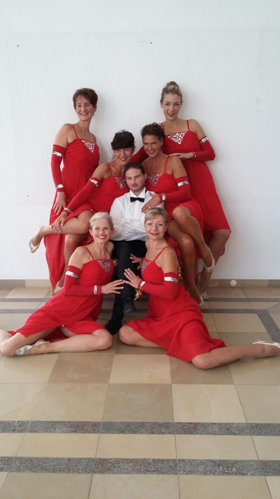 Gruppenszene mit sieben Personen in roten Kleidern, um einen Mann in formeller Kleidung, in einem hellen Raum.