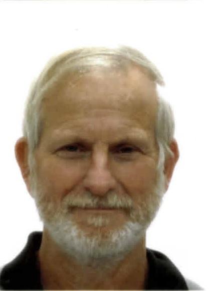 Ein älterer Mann mit grauem Haar und Bart sitzt vor einem neutralen Hintergrund und lächelt direkt in die Kamera.