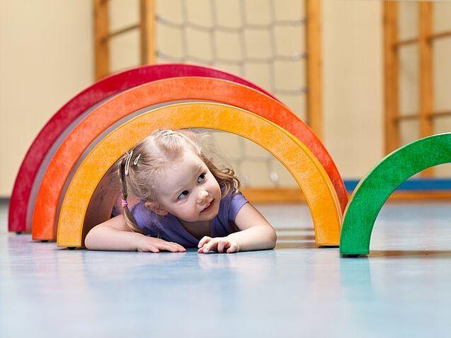 M&auml;dchen kriecht unter bunten, halbkreisf&ouml;rmigen Spielb&ouml;gen in einem Spielraum.