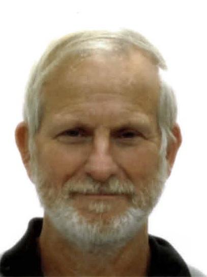 Ein älterer Mann mit grauem Haar und Bart sitzt vor einem neutralen Hintergrund und lächelt direkt in die Kamera.