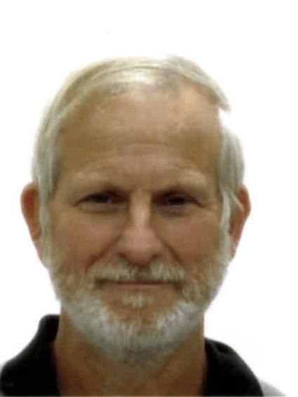 Ein älterer Mann mit grauem Haar und Bart sitzt vor einem neutralen Hintergrund und lächelt direkt in die Kamera.