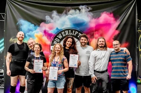 Gruppe von acht Menschen mit Urkunden vor einem bunten Hintergrund bei einer Powerlifting-Veranstaltung.
