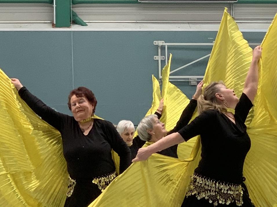 Tanzgruppe in schwarzen Kleidern mit goldenen Accessoires, die gelbe Stoffe lebhaft in einer Sporthalle schwenkt.