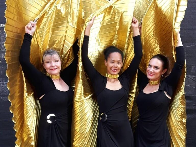 Drei Frauen in schwarzen Outfits halten goldene, gefaltete Stoffelemente &uuml;ber ihren K&ouml;pfen vor einer dunklen Hintergrundb&uuml;hne.