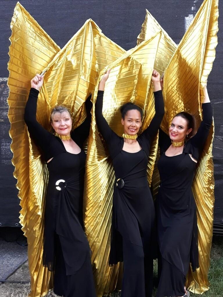 Drei Frauen in schwarzen Outfits halten goldene, gefaltete Stoffelemente über ihren Köpfen vor einer dunklen Hintergrundbühne.
