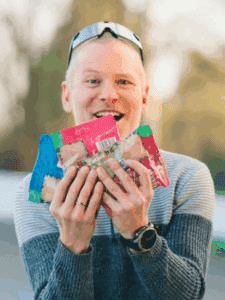 Mann in Sportkleidung hält mehrere Packungen Snacks und lächelt erfreut in die Kamera. Sonnenbrille auf dem Kopf.