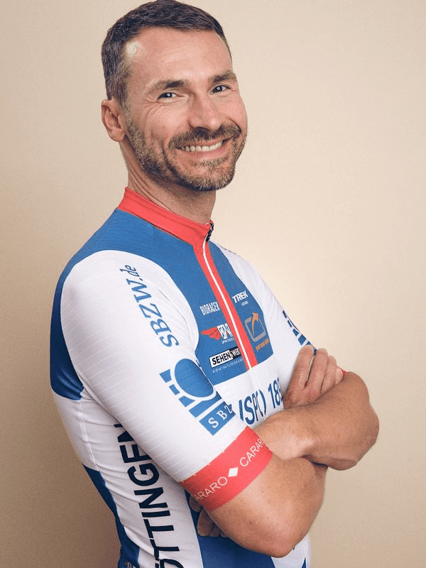 Lächelnder Radfahrer in hautenger, blauer und weißer Trikotausführung, mit verschränkten Armen vor neutralem Hintergrund.