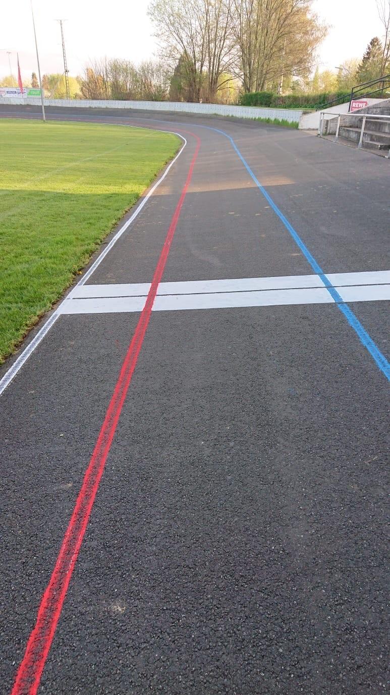 Kurve einer Radrennbahn mit rot und blau markierten Linien sowie einer weißen Ziellinie auf asphaltiertem Untergrund.