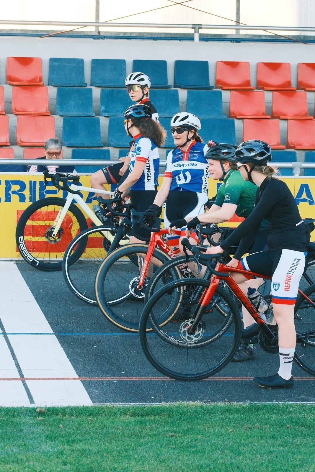 Sechs Radfahrerinnen in Rennkleidung stehen neben ihren Fahrr&auml;dern auf einem Radrennbahn-Startplatz.