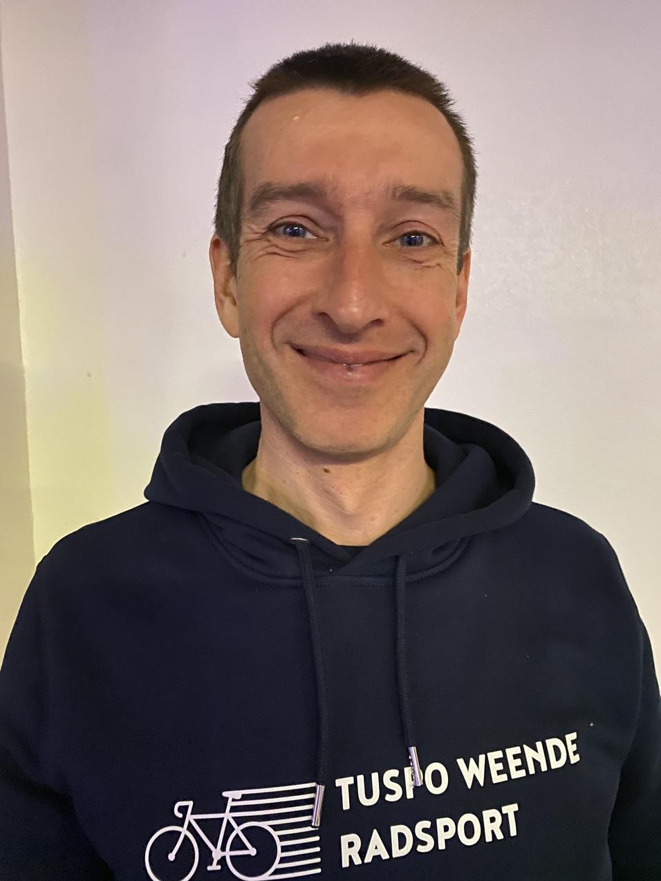 Portrait eines lächelnden Mannes in einem dunkelblauen Sweatshirt mit der Aufschrift „TUSPO WEENDE RADSPORT“.