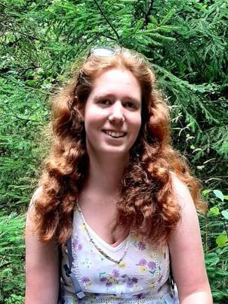 Frau mit lockigem, rotem Haar sitzt auf einem umgestürzten Baum im Wald, umgeben von grünen Pflanzen.