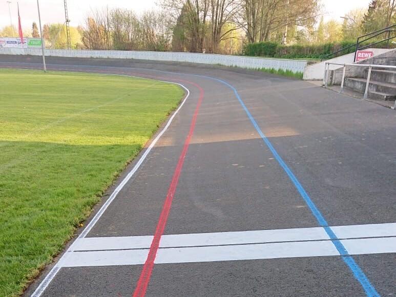 Kurve einer Radrennbahn mit rot und blau markierten Linien sowie einer wei&szlig;en Ziellinie auf asphaltiertem Untergrund.