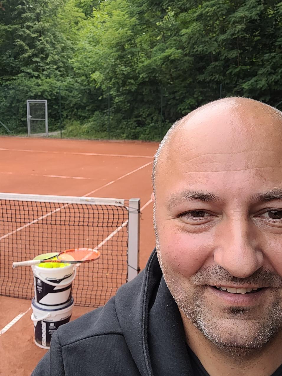 Selbstportrait eines Mannes mit Tennisplatz und Schläger im Hintergrund, umgeben von Bäumen.