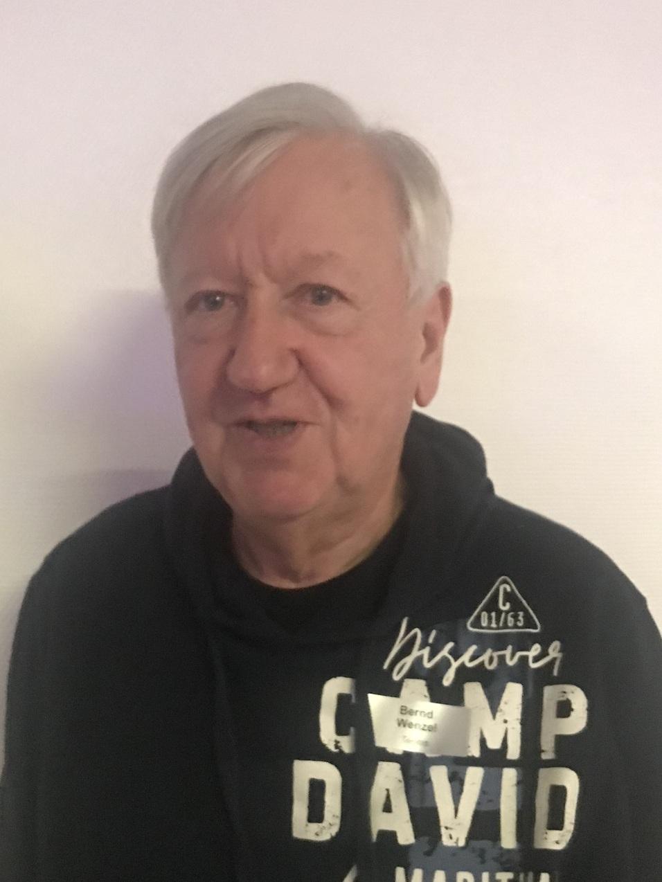 Älterer Mann mit grauen Haaren steht vor einer weißen Wand, trägt einen schwarzen Pullover mit Aufdruck und ein Namensschild.