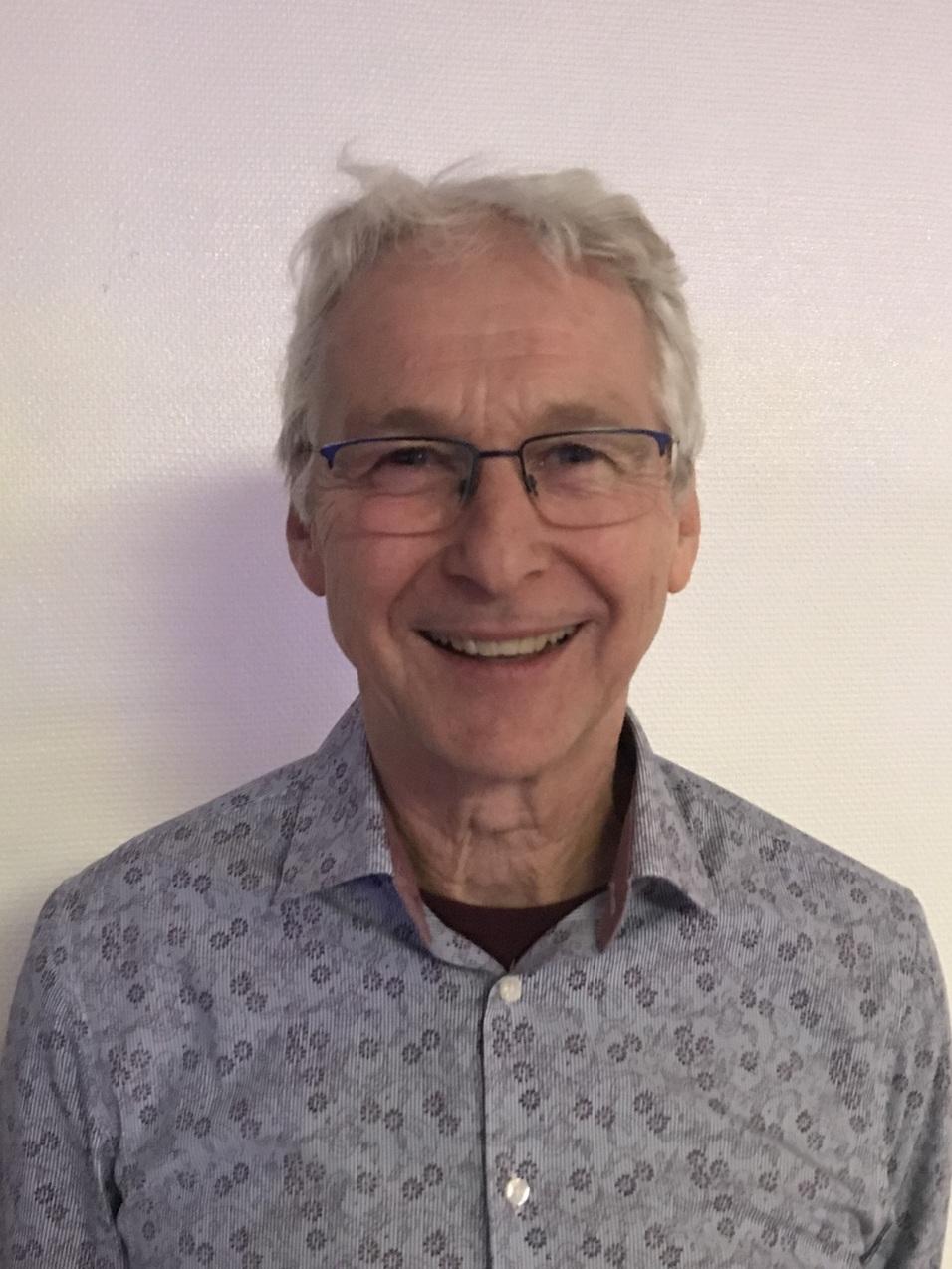 Portrait eines lächelnden älteren Mannes mit Brille und Hemd mit floralem Muster vor einer hellen Wand.