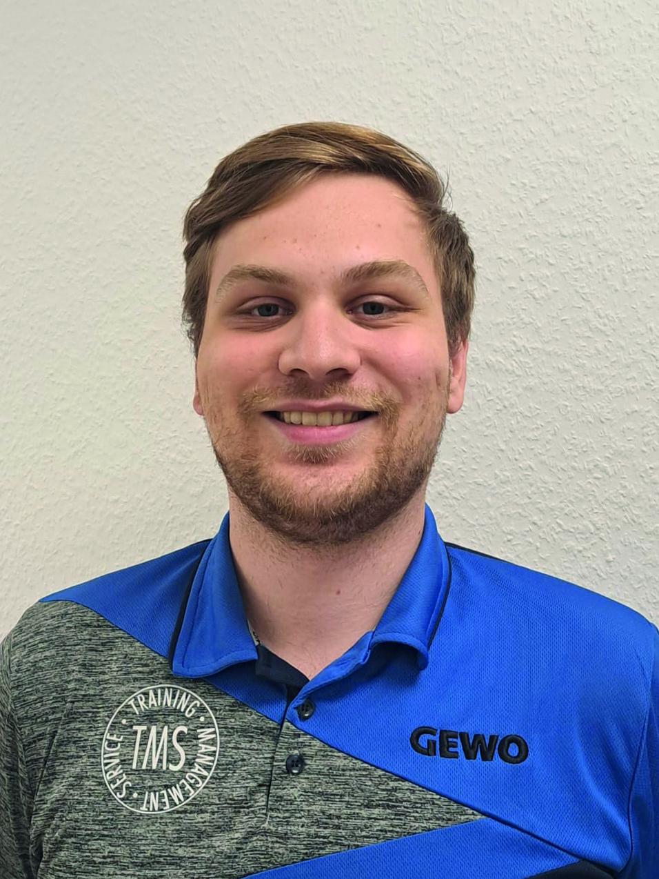 Lächelnder junger Mann in Sportbekleidung mit dem Logo „GEWO“, vor grauer Wand stehend.