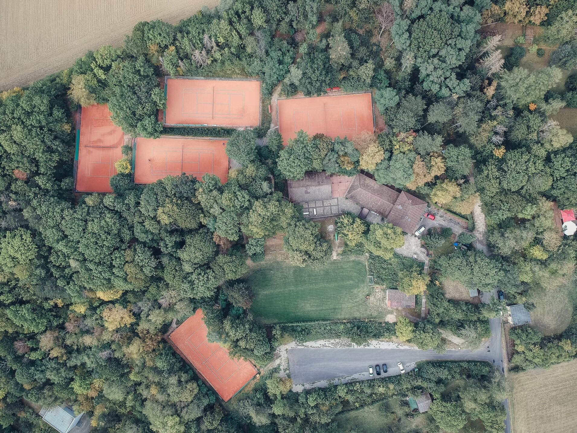 Luftaufnahme eines Tennisplatzes, umgeben von B&auml;umen, mit einer nahegelegenen Wohnstruktur und einer Wiese.