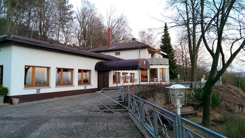 Ansicht eines Gebäudes mit Veranda, großen Fenstern und Terrasse, umgeben von Bäumen und einer Grünfläche.