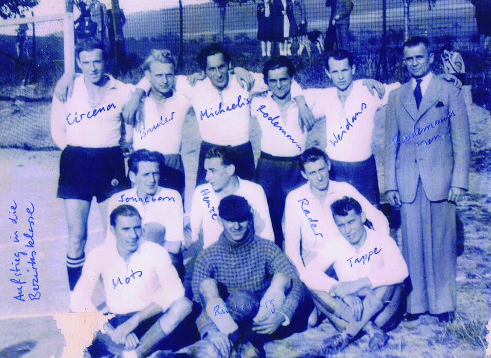Schwarz-weiß Foto einer Fußballmannschaft mit Namen der Spieler in blauer Schrift, auf einem Sportplatz vor einem Zaun.