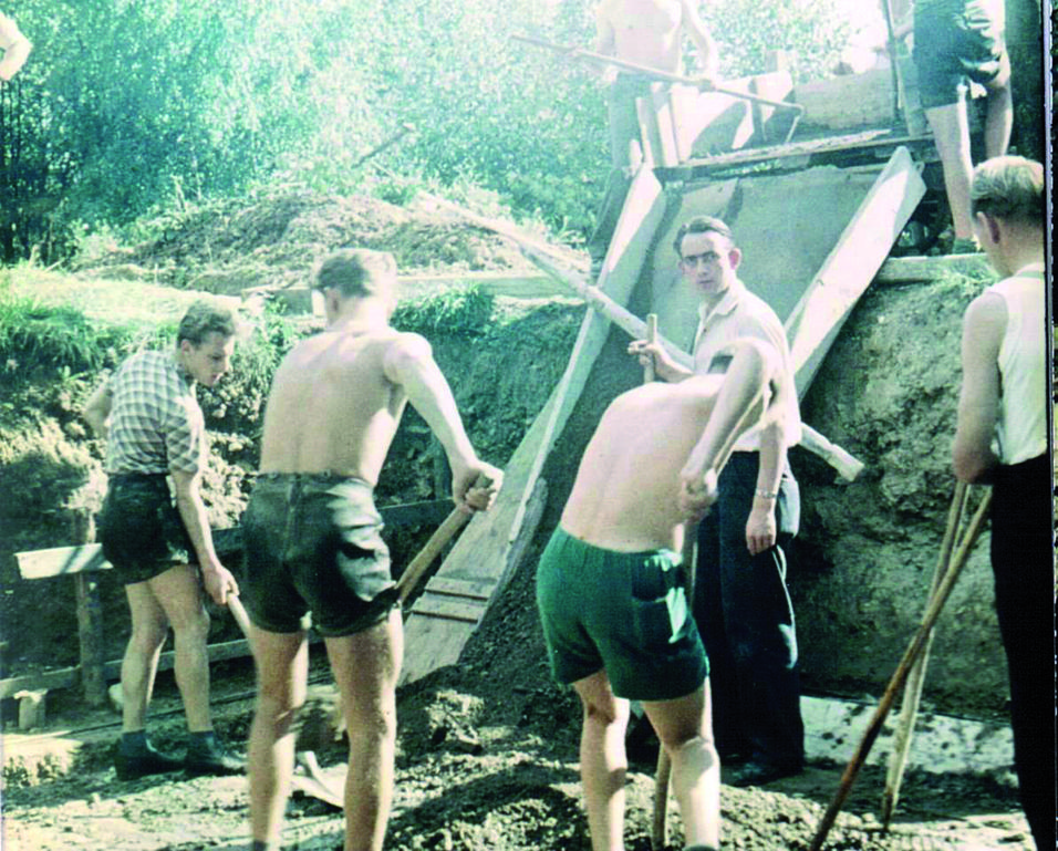 Gruppeneinteilung beim Bau: Mehrere Männer arbeiten mit Schaufeln am Hang, während ein weiterer Zuschauer an der Rampe steht.